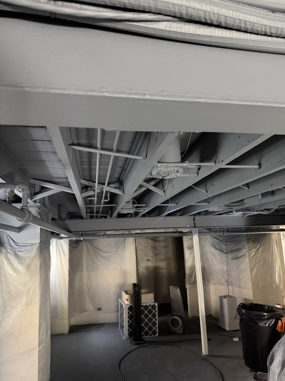 Basement ceiling joists, pipes and beam fully primed in gray, walls masked in plastic